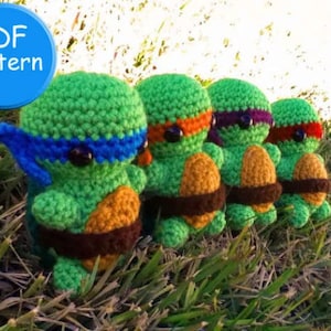 May include: Four crocheted Teenage Mutant Ninja Turtles, each with a different colored bandana. The turtles are green with brown shells and orange undershells. The turtles are standing on a bed of green grass.
