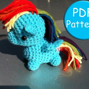 Könnte beinhalten: Ein gehäkeltes Regenbogen-Pony-Amigurumi-Spielzeug mit einem blauen Körper, einer roten, gelben, orangen, grünen und violetten Garnmähne und einem Schwanz. Das Spielzeug sitzt auf einer grauen Oberfläche. Der Text "PDF Pattern!" befindet sich auf einem blauen Kreis.
