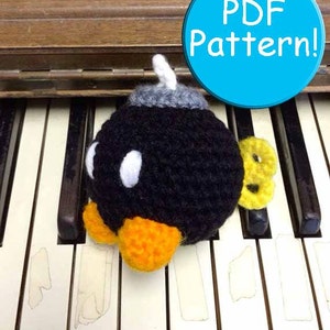 Könnte beinhalten: Ein gehäkeltes schwarzes Bomben-Plüschtier mit gelben und orangefarbenen Akzenten, das auf einer weißen und schwarzen Klaviertastatur sitzt. Der Text "PDF Pattern!" befindet sich auf einem blauen Kreis im Hintergrund.