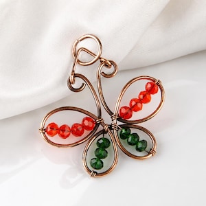 May include: A butterfly-shaped pendant crafted from copper-colored wire. The wings are formed by wire loops containing faceted beads in vibrant red and deep green colors. A circular wire loop at the top serves as a bail for hanging the piece.