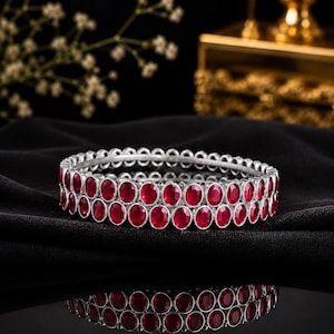 Puede incluir: Un brazalete de doble fila que presenta una serie de piedras ovaladas de color rojo intenso engastadas en un marco de metal plateado. Las piedras están dispuestas en dos líneas paralelas, creando un diseño circular continuo con acabado pulido.