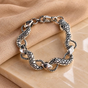 Könnte beinhalten: Ein silberfarbenes Metallarmband mit einem sich wiederholenden Schlangenmotiv und detaillierten Schuppenstrukturen. Das Design besteht aus miteinander verbundenen Schlangensegmenten, die eine kreisförmige Kette bilden und mit einem Karabinerverschluss aus Metall gesichert sind.