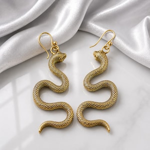 May include: A pair of gold-toned metal earrings shaped like coiled snakes with textured scales. Each earring features a curved hook for attachment, with the snake body winding in an S-shape, ending in a tapered tail. The metal has a polished finish.