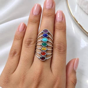 Op de afbeelding: Een zilveren ring met meerdere banden bestaande uit zeven op elkaar gestapelde, dunne metalen draden. Elke draad is versierd met een kleine, ovale cabochon edelsteen in een verticale opstelling, in de kleuren paars, blauw, turquoise, groen, geel, oranje en rood.