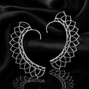 May include: A pair of silver-toned ear cuffs featuring an intricate, openwork filigree design that resembles a series of connected petal shapes. The curved, crescent-like structure follows the contour of the ear, creating a delicate, lace-like appearance.