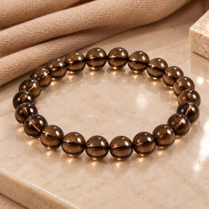 May include: A circular bracelet composed of numerous smooth, translucent, dark brown smoky quartz beads strung together. The beads are uniform in size and shape, featuring a polished, reflective surface that catches the light, creating a glossy appearance.