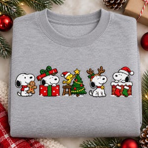 Peut inclure: Un sweat-shirt gris chiné à col rond orné d'une impression graphique de Snoopy et Woodstock dans diverses scènes de Noël, notamment avec un bonhomme en pain d'épice, sortant d'un cadeau, décorant un sapin et portant des bois de renne.
