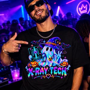 May include: A black t-shirt featuring a colorful graphic of a ghost wearing a witch hat and a stethoscope, holding an X-ray image. The design includes two jack-o'-lanterns, bats, and the text X-RAY TECH in a vibrant, dripping neon font style.