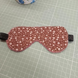 Può includere: Una mascherina per dormire in tessuto marrone con motivo floreale bianco e bordo rosa testurizzato. La mascherina presenta fasce laterali elastiche nere ed è posizionata su un tappetino da taglio verde con griglia bianca e misure in cm.