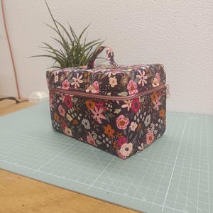 Pode incluir: Um estojo de armazenamento retangular em tecido com fundo roxo escuro e um padrão floral multicolorido de flores cor-de-rosa, laranja e brancas. O estojo inclui uma pega superior, um fecho de correr e uma forma estruturada e quadrada.