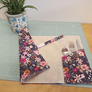 Op de afbeelding: Een stoffen bestekrol met een donkerblauwe achtergrond en een veelkleurig bloemenpatroon in roze, oranje en witte tinten. De binnenkant is van lichtbeige stof met vakjes waarin een zilverkleurige metalen lepel, vork en mes zitten.