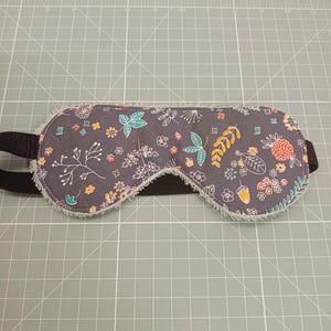 Op de afbeelding: Een slaapmasker met een donkergrijze stoffen basis en een herhalend patroon van kleine witte, oranje en turquoise bloemen, bladeren en eikels. Het masker is voorzien van een lichte, pluizige rand en een zwarte elastische band voor bevestiging.