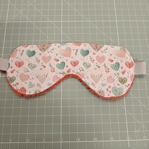 Könnte beinhalten: Eine Schlafmaske mit weißer Stoffvorderseite, bedruckt mit einem wiederkehrenden Muster aus rosa und blaugrünen Herzen sowie kleinen Blumenmotiven. Die Maske hat einen strukturierten orangefarbenen Rand und ein hellgraues elastisches Band an den Seiten.