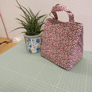 Pode incluir: Uma pequena bolsa de tecido com um padrão floral denso em tons de vermelho, rosa e verde sobre um fundo branco. A bolsa possui duas alças curtas e um fecho de botão de pressão no centro superior, feita de um material têxtil leve.