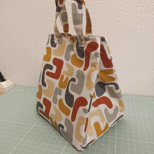 Pode incluir: Uma bolsa de tecido com um padrão abstrato repetitivo de formas curvas em tons de laranja queimado, amarelo mostarda e cinza sobre um fundo creme. A bolsa tem duas alças de tecido a condizer e um fecho de botão de pressão no topo.