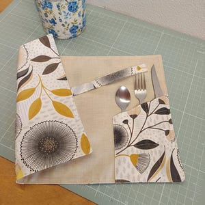 Peut inclure: Un set de table en tissu avec un centre beige et une superposition blanche ornée d'un motif floral et de feuilles jaune moutarde, gris foncé et marron. Le set comprend une poche intégrée contenant une cuillère, une fourchette et un couteau.