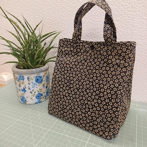 Peut inclure: Un sac fourre-tout en tissu bleu marine foncé orné d'un motif répétitif de petites fleurs claires à quatre pétales. Le sac est doté de deux anses en tissu assorties et d'une fermeture à bouton-pression unique au centre de l'ouverture.