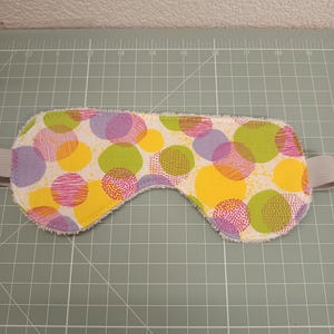 Op de afbeelding: Een slaapmasker met een witte stoffen basis en een patroon van overlappende cirkels in geel, limoengroen en lavendel. De cirkels bevatten verschillende texturen zoals stippen en fijne lijntjes. Het masker heeft een witte badstof rand en grijze elastische banden.