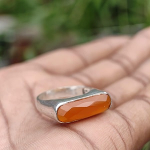 May include: A silver ring featuring a long, rectangular, faceted orange gemstone set horizontally. The stone has a smooth, polished surface with multiple geometric facets that catch the light, held within a simple, solid silver band frame.