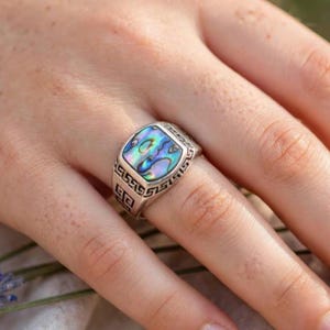 May include: A silver ring featuring a square-shaped abalone shell inlay with iridescent blue, green, and purple tones. The band is crafted with a detailed Greek key pattern engraved along the sides of the setting, framing the central stone.