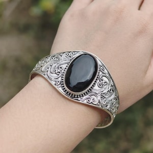 May include: A silver-toned cuff bracelet featuring an oval-shaped, smooth black stone set in the center. The metal band is decorated with intricate, swirling filigree patterns that extend across the entire surface of the cuff.