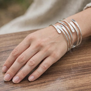 Puede incluir: Una pulsera tipo brazalete de tono plateado con un diseño en espiral de varias capas que se envuelve alrededor de la muñeca. La pulsera está compuesta por varias bandas de metal liso y pulido que se estrechan en los extremos, creando una forma fluida.