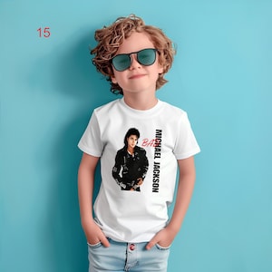 May include: A white short-sleeved t-shirt featuring a graphic print of Michael Jackson in a black leather jacket with the text BAD in red and MICHAEL JACKSON in black. The shirt is worn with light blue denim jeans and dark sunglasses.