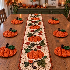 May include: A long, cream-colored crocheted table runner featuring a repeating pattern of orange pumpkins with green leaves and small red accents, accompanied by six matching orange pumpkin-shaped crocheted coasters arranged on a wooden table.