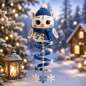 Op de afbeelding: Een gehaakt wit uilornament met grote zwarte ogen, een gele snavel, een blauwe gebreide muts en een bijpassende blauwe sjaal. Onder de uil hangt een spiraalvormige staart met blauw en wit garen, houten kralen en drie bungelende sneeuwvlokken.