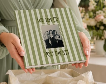 May include: A square photo album with a sage green and white vertical striped cover. The front features a central black and white photograph of three people framed by a scalloped border, with the text WE LOVED YOU FIRST! printed in green.