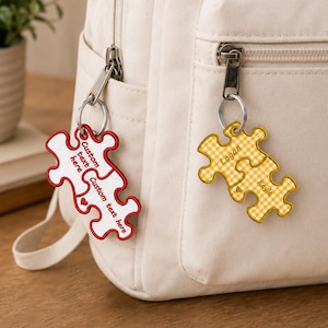 Op de afbeelding: Twee sets puzzelstuk-sleutelhangers bevestigd aan een crèmekleurige rugzak. De ene set is wit met rode randen en de tekst Custom text here. De andere set is geel met een wit gingham-patroon en de namen Logan en Kayle erop gedrukt.