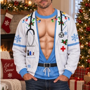 Puede incluir: Una sudadera blanca de manga larga con un diseño estampado de bata de médico sobre uniforme azul con un torso musculoso. El diseño incluye un estetoscopio, bolígrafos, una cruz roja, acebo, copos de nieve y luces navideñas de colores.