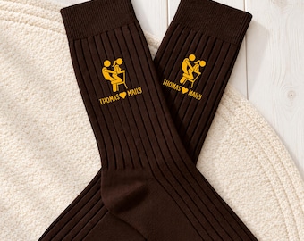 May include: A pair of dark brown ribbed socks featuring a yellow graphic of two figures on a chair and the text "THOMAS HEART MAILY" printed in yellow on the upper section of each sock, positioned in a crossed arrangement.