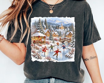May include: A charcoal gray short-sleeve t-shirt featuring a rectangular graphic print of a winter scene. The illustration depicts children ice skating on a frozen pond near a gazebo, a stone bridge, and snow-covered houses with mountains.