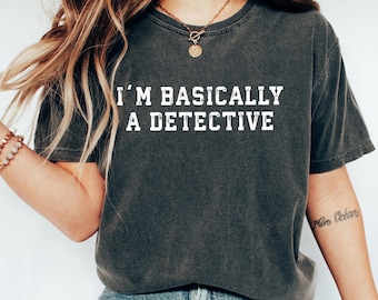 May include: A charcoal gray short-sleeve t-shirt featuring the white distressed-style text I'M BASICALLY A DETECTIVE printed across the chest. The shirt has a crew neckline and a relaxed fit, paired with blue denim jeans.