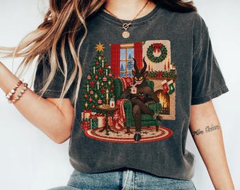 May include: A charcoal gray t-shirt featuring a graphic print of a horned, furry creature sitting in a green armchair by a fireplace and a decorated Christmas tree. The scene includes a wreath, stockings, and gifts, with the creature holding a mug.