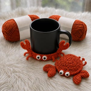 May include: A matte black ceramic mug sits inside a crocheted orange crab coaster with white and black eyes. A separate matching crocheted orange crab coaster lies next to the mug. Two skeins of orange yarn with white labels are positioned behind the mug.