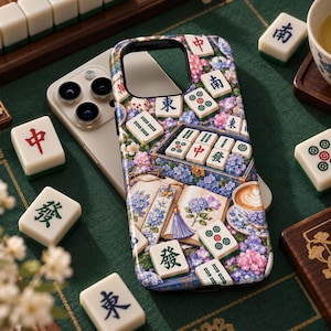 May include: A smartphone case featuring a detailed print of scattered Mahjong tiles, an open book with floral illustrations, a blue tassel bookmark, and a cup of coffee, all surrounded by clusters of blue, purple, and pink hydrangea flowers.