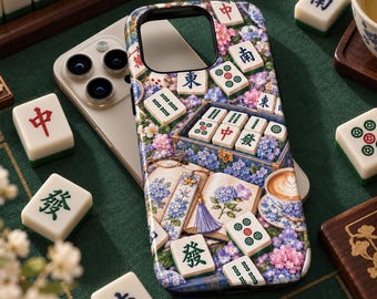 May include: A smartphone case featuring a detailed print of scattered Mahjong tiles, an open book with floral illustrations, a blue tassel bookmark, and a cup of coffee, all surrounded by clusters of blue, purple, and pink hydrangea flowers.