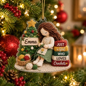 May include: A clear acrylic Christmas ornament featuring a crocheted-style illustration of a brown-haired figure crocheting next to a decorated tree. The tree has a sign reading Emma. A stack of books reads JUST a Girl WHO LOVES Crochets.