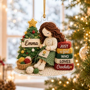 May include: A clear acrylic Christmas ornament featuring a crocheted-style illustration of a person with brown hair crocheting next to a decorated Christmas tree. The tree has a sign reading Emma. A stack of books reads Just a Girl Who Loves Crochets.