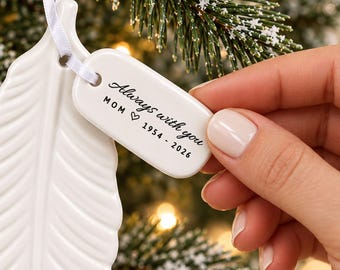 Op de afbeelding: Een wit keramieken ornament in de vorm van een veer met een rechthoekig label bevestigd aan een wit lint. Het label is bedrukt met de zwarte tekst Always with you, MOM, een hartje en de jaartallen 1954 - 2026.