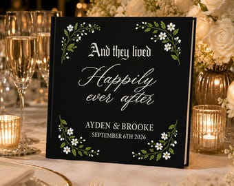 May include: A square black guest book featuring white floral vine illustrations in the corners and the text And they lived Happily ever after, followed by the names AYDEN & BROOKE and the date SEPTEMBER 6TH 2026 in white lettering.