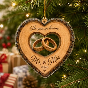 May include: A heart-shaped wooden ornament with bark edges, featuring two interlocking rings in the center. The text reads: The year we became, Lucas, Sophia, Mr. & Mrs., and 2026 with a small heart icon. It hangs from a gold string.