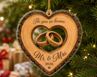 May include: A heart-shaped wooden ornament with bark edges, featuring two interlocking rings in the center. The text reads: The year we became, Lucas, Sophia, Mr. & Mrs., and 2026 with a small heart icon. It hangs from a gold string.