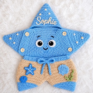 May include: A blue crochet star-shaped hooded garment with the name Sophie embroidered on the hood. The piece features four wooden buttons, large white and black eyes, and tan shorts adorned with a blue starfish, two blue shells, and green seaweed.