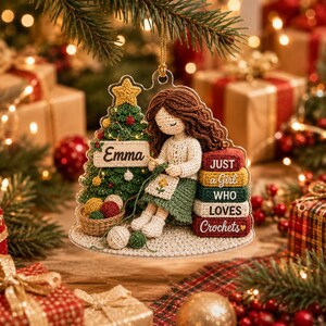 May include: A clear acrylic Christmas ornament featuring a crocheted-style illustration of a person with brown hair crafting next to a decorated tree. The tree has a sign reading Emma, and a stack of books reads JUST a Girl WHO LOVES Crochets.