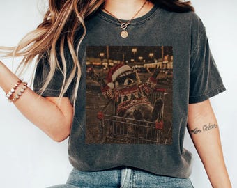 May include: A charcoal gray short-sleeve t-shirt featuring a graphic print of a raccoon wearing a red and white Santa hat, dark sunglasses, and a patterned holiday sweater while sitting inside a metal shopping cart at night.