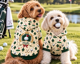May include: Two dogs wearing cream-colored hoodies with a golf-themed pattern of balls, flags, and clubs. The larger dog's hoodie features a green crest with a crown and the name Bella. The smaller dog holds a white golf ball in its mouth.