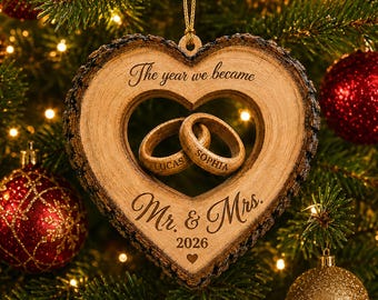 May include: A heart-shaped wooden ornament with bark edges, featuring two interlocking wooden rings engraved with the names Lucas and Sophia. The ornament is inscribed with the text The year we became Mr. & Mrs. 2026 and a small heart icon.